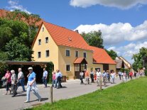 Spazierende Menschen vor einem gelben Haus bei schönem Wetter und Wolken am Himmel. 