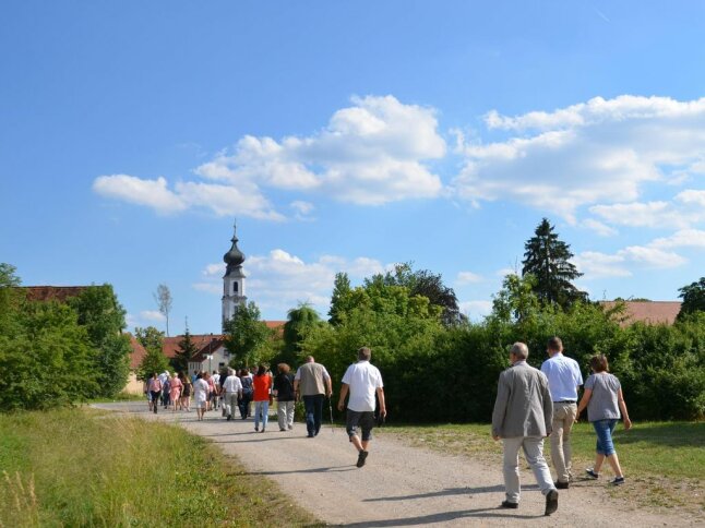 Menschen spazieren in Richtung eines eingegrünten Dorfes. Der große Kirchturm ragt in einen weiß-blauen Himmel